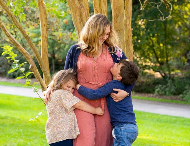 Woman hugging two children outdoors with trees and grass in the background